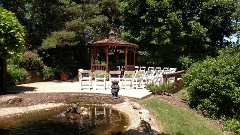 Outdoor wedding ceremony setup by a pond, featuring a wooden gazebo with white chairs arranged in front. Lush greenery surrounds the area, creating a serene and natural atmosphere.