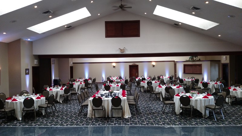 Large banquet hall set with round tables covered in white linens and red napkins, arranged for an event with chairs neatly positioned, skylights above, and neutral walls surrounding the space.