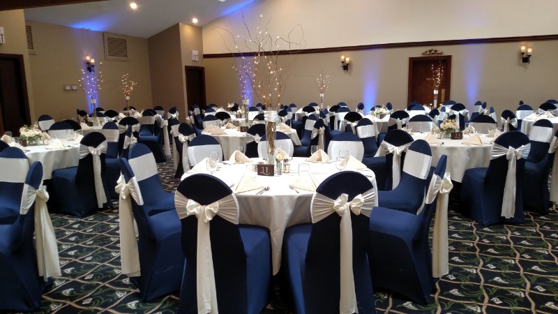 Banquet hall set up with round tables covered in white tablecloths and blue chairs, each adorned with a white satin chair sash. Soft blue lighting illuminates the room.